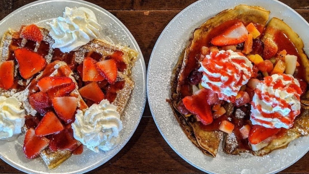 A plate of waffles and a plate of crepes both with strawberries, whipped cream and icing sugar on them